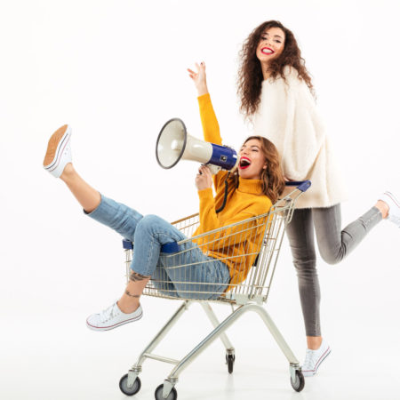 Two happy girls in sweaters having fun with shopping trolley and megaphone over white background